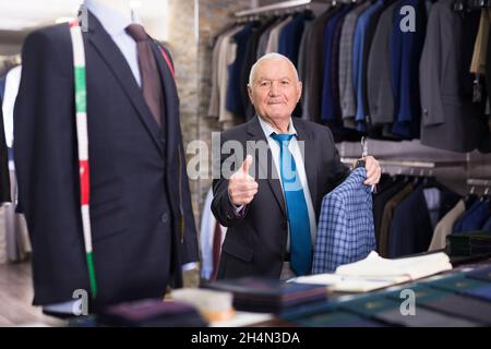 Felice anziano che sceglie la giacca alla moda in negozio di abbigliamento e mostra pollice in su Foto Stock