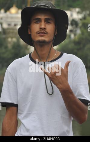 Ritratto di un giovane indiano bello osservare che fa segno shaka con la sua mano con cappello e maglietta bianca e guardando la macchina fotografica Foto Stock