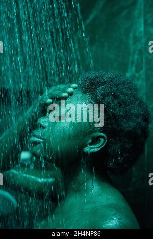 Donna africana in piedi sotto l'acqua e lavarsi il viso e i capelli in bagno Foto Stock
