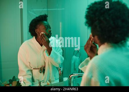 Donna africana in accappatoio guardando lo specchio mentre si lava il suo trucco con spugna in bagno Foto Stock