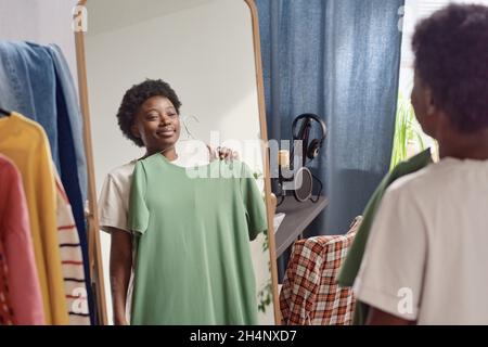 Giovane donna africana in piedi davanti allo specchio e provare sul nuovo vestito mentre si alza in piedi nella stanza Foto Stock