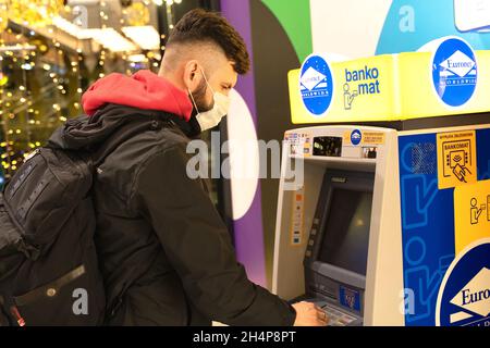 POLONIA, TORUN - 5 dicembre 2020: L'uomo ritira contanti vicino a ATM. Il ragazzo usa il credito per soldi. Persona in stile casual indossare maschera protettiva sul suo viso i Foto Stock