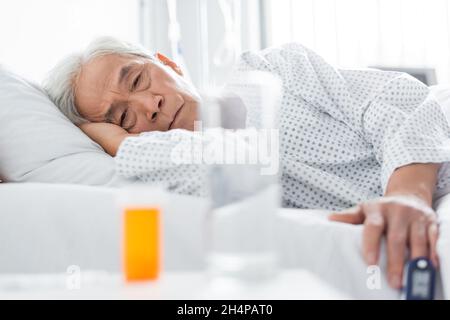 Paziente asiatico anziano con pulsossimetro che guarda pillole e acqua in clinica Foto Stock