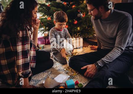 Ragazzo che apre il suo regalo di Natale. Famiglia seduta sul pavimento da albero di Natale unwrapping regali di Natale. Foto Stock