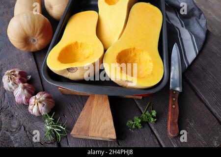 Zucca di zucca di zucca al burro dimezzata sul tagliere di legno. Coltello e asciugamano da cucina su sfondo scuro Foto Stock