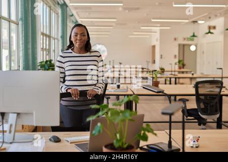 Fidata giovane donna d'affari africana in piedi alla sua scrivania in un ufficio Foto Stock