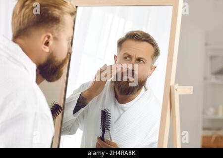 Uomo preoccupato che guarda le rughe sul viso nello specchio durante la routine di cura della pelle Foto Stock