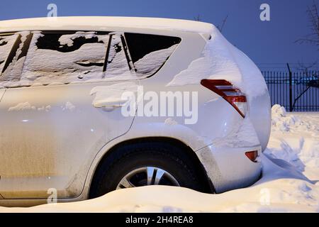 Auto bianca coperta di neve di notte. SUV parcheggiato nel cortile. Città dopo la tempesta di neve. Vista laterale. Foto Stock