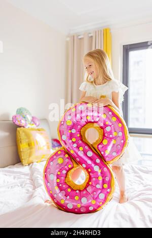 Scatto interno di grazioso capretto adorabile della ragazza gioioso che celebra il compleanno di otto anni con i palloncini luminosi e colorati con le parole di iscrizione soggiorno Foto Stock