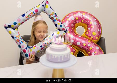 Felice piccola ragazza caucasica con emozione positiva sul volto festeggiando il compleanno di otto anni a casa. Candido lifestyle ritratto di Kid soffiando Foto Stock