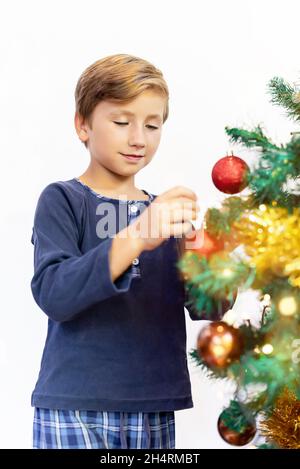 Ragazzo felice in pigiama decorando l'albero di Natale, mettendo palle di Natale su di esso in una fredda mattina di inverno Foto Stock
