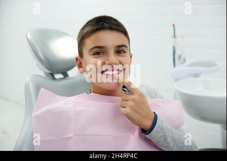 Il ragazzo felice tiene il dito vicino alla bocca, guarda la macchina fotografica, sorride con un bel sorriso toothy dopo aver ricevuto il trattamento dentale in clinica odontoiatria. Orale Foto Stock