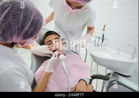 Ragazzo di scuola seduto sulla sedia del dentista che riceve il trattamento medico della sua cavità orale da parte del dentista pediatra e il suo assistente nella moderna cli odontoiatria Foto Stock