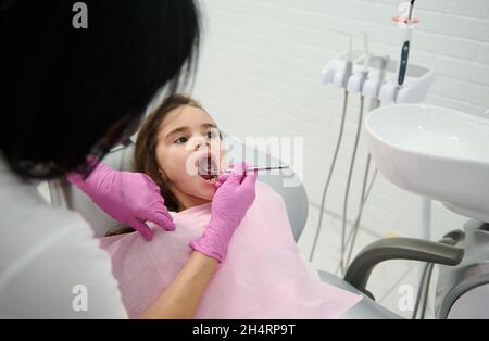 Adorabile bambina seduta nella sedia del dentista con la bocca aperta, mentre il dentista pediatrico esamina la sua cavità orale utilizzando un acciaio inossidabile d Foto Stock