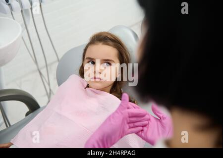 Bambina seduta in sedia dentista, ascoltando un igienista pediatrico che le insegnava l'igiene orale e la prevenzione del decadimento dei denti del bambino durante la denta Foto Stock