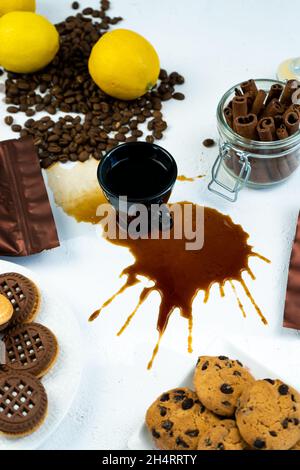 Caffè versato da una tazza nera su un tavolo bianco. Chicchi di caffè, cannella e biscotti. Prima colazione in cucina. Foto Stock