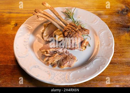 Costolette di agnello al forno con patate e rosmarino in piatto bianco su rustico tavolo di legno, luce calda, vista dall'alto Foto Stock