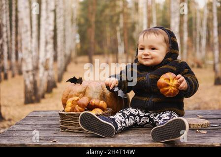 Bambino di 10 mesi si siede con zucca e bun su tavola di legno nella foresta d'autunno. Concetto di vacanza di Halloween. Foto Stock