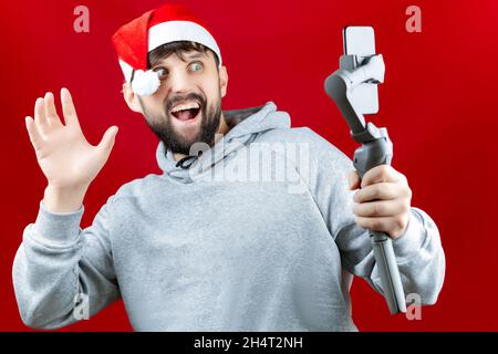 Un uomo allegro in un cappello rosso di Babbo Natale tiene in mano uno stabilizzatore elettronico su cui il telefono di un uomo prende un selfie Foto Stock