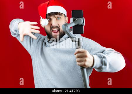 Un uomo allegro in un cappello rosso di Babbo Natale tiene in mano uno stabilizzatore elettronico su cui il telefono di un uomo prende un selfie Foto Stock