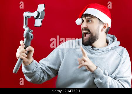 Un uomo divertente in un cappello rosso di Babbo Natale con uno stabilizzatore nelle sue mani grimaces alla fotocamera dello smartphone Foto Stock