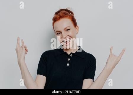 Giovane donna rossa sicura che fa segno di rock and roll mentre si alza isolato su sfondo grigio Foto Stock