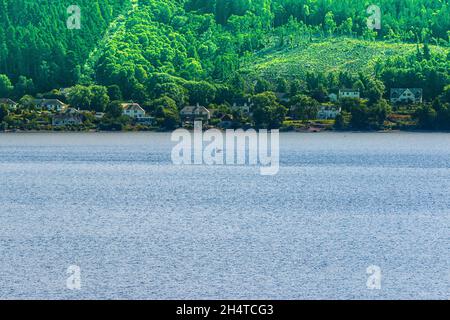 Vista su un lago in Scozia. Loch Ness di giorno in barca. Case sulla riva con foresta e montagne sullo sfondo in estate. Alberi verdi sul Foto Stock