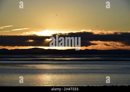 Il sole tramonta verso il basso rivelando riflessi d'acqua scintillanti e nuvole scure sopra l'estuario del fiume Mersey. Foto Stock