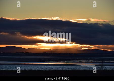 Il sole tramonta verso il basso rivelando riflessi d'acqua scintillanti e nuvole scure sopra l'estuario del fiume Mersey. Foto Stock