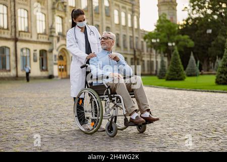 Scatto a tutta lunghezza del paziente anziano handicappato in sedia a rotelle sorridente al giovane infermiere in maschera protettiva aiutandolo all'aperto Foto Stock