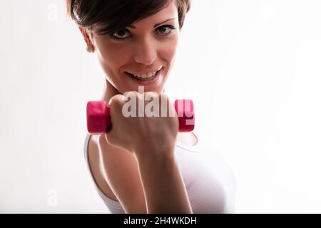 Determinata giovane donna che si sta allenando con pesi che sollevano un manubbell in mano in primo piano con il fuoco al suo viso Foto Stock