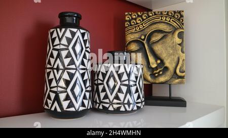 Decorazioni per la casa con vasi in ceramica bianca e nera e un Buddha dorato faccia figurine contro parete rossa. Foto Stock