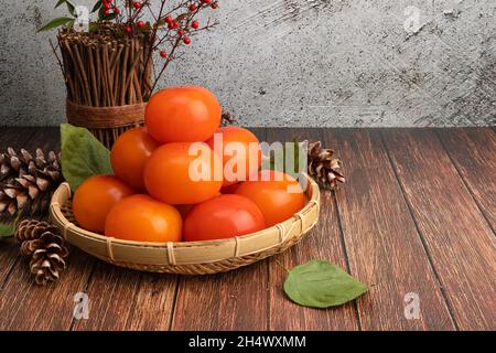 Persimmons maturi in un cesto di bambù. Sfondo tavola in legno con un set di persimmons a sinistra. Foto Stock