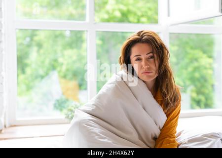 la donna di 40 anni è malata. Si siede sul letto coperto da una coperta e guarda tristemente la macchina fotografica. Foto Stock