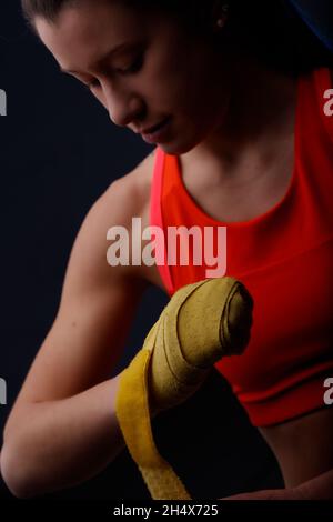 Giovane e forte donna in rosso di allenamento top che avvolge i suoi pugni con le mani gialle avvolge prima di boxe di allenamento. Ritratto in primo piano contro retro nero scuro Foto Stock