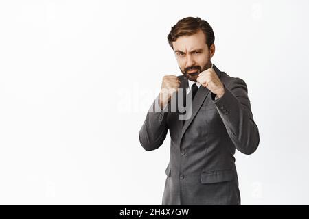 Un uomo d'affari serio e bello stringe pugni, guardando la macchina fotografica osare, combattendo, pugilato con qualcuno, in piedi su sfondo bianco Foto Stock
