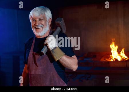 Fabbro caucasico invecchiato in grembiule protettivo e guanti sorridenti sulla fotocamera mentre tiene il martello sulla spalla. Ritratto di buon artigiano maturo con g Foto Stock