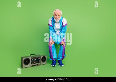 Foto di cool funky uomo anziano indossare tuta sportiva ballare ascolto boombox sorridente isolato sfondo di colore verde. Foto Stock