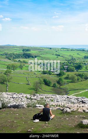 Giovane uomo viaggio da solo, vista posteriore in estate di un uomo seduto vicino al bordo della scogliera del marciapiede calcareo a Malham Cove nel Yorkshire Dales, Inghilterra Foto Stock