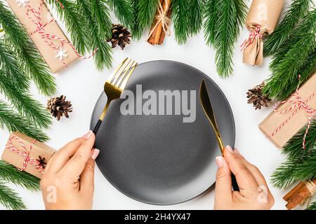 Decorazioni di Natale festive tavolo. Le mani femminili tengono le posate dorate sopra la piastra vuota nera Foto Stock