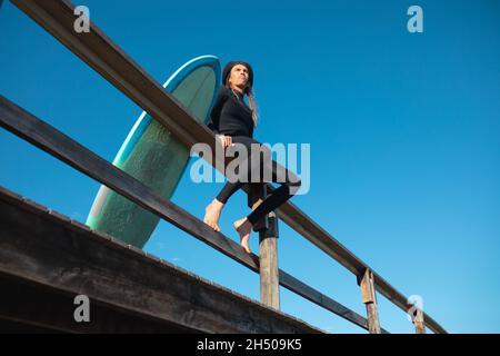 Vista ad angolo basso dell'uomo seduto con tavola da surf sulla ringhiera contro il cielo blu chiaro con spazio copia Foto Stock