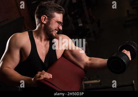 Un bell'uomo in occhiali con una barba sta facendo un esercizio per bicipite con un barbell. Il ragazzo giovane è impegnato in bodybuilding. Il formatore in Foto Stock