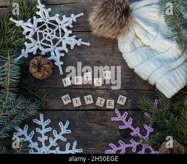 ornamenti in fiocco di neve a crochet su terreno di legno con slogan invernali e ramo di abete e coni e un cappello a maglia Foto Stock