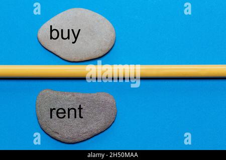 Le parole buy e rent sono scritte su pietre naturali lisce separate da una matita gialla. Lo sfondo è isolato in blu e ha molto spazio Foto Stock