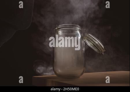 Vaso di vetro con coperchio riempito di fumo su sfondo nero. Nebbia in un contenitore trasparente sul tavolo. Foto Stock