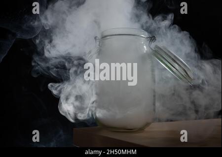 Vaso di vetro con coperchio riempito di fumo su sfondo nero. Nebbia in un contenitore trasparente sul tavolo. Foto Stock