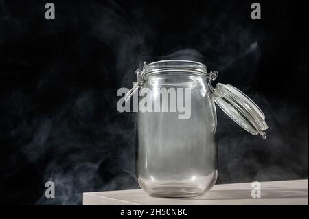 Vaso di vetro con coperchio riempito di fumo su sfondo nero. Nebbia in un contenitore trasparente sul tavolo. Foto Stock