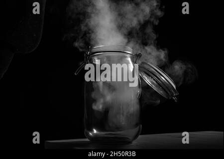 Vaso di vetro con coperchio riempito di fumo su sfondo nero. Nebbia in un contenitore trasparente sul tavolo. Foto Stock