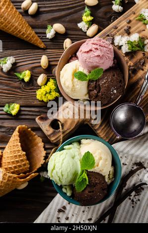 Diversi scoop di cioccolato al pistacchio alla vaniglia e palline di icecream alla fragola in ciotole di argilla su tavolo da cucina in legno. Disposizione piatta Foto Stock