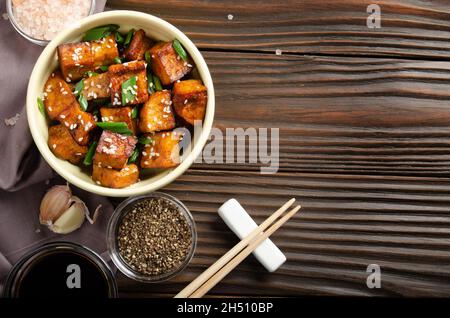 Vista piana a disposizione al tofu fritto di mescolare cubetti con erba cipollina in piatto di argilla su tavolo da cucina in legno con erba cipollina cipollina pepe aglio e salsa di soia a parte Foto Stock